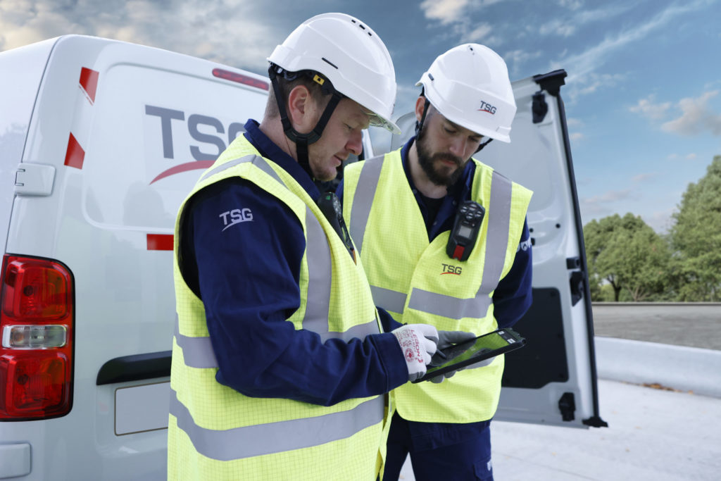 Twee techniekers van TSG met een tablet in de hand. Ze bekijken een schema met op de achtergrond een bestelwagen van TSG.