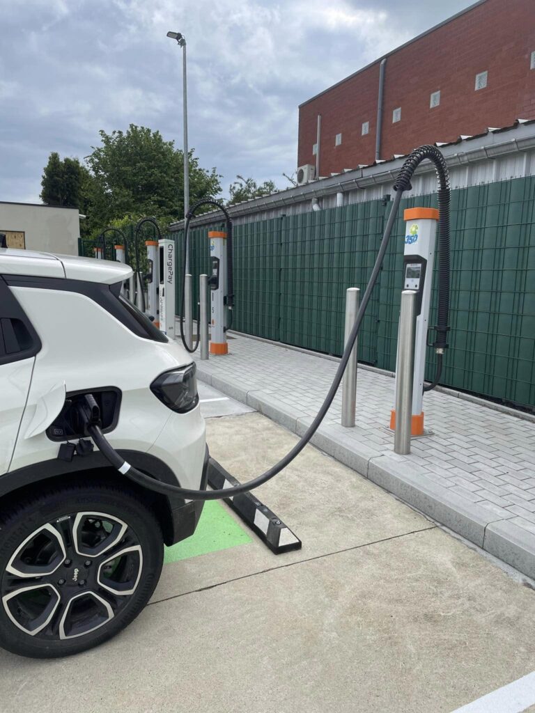 Een witte wagen laadt op aan een Kempower-laadsatelliet op het laadplein van Station Interzonings. Op de achtergrond zie je nog enkele Kempower-satellieten en de ChargePay terminal.